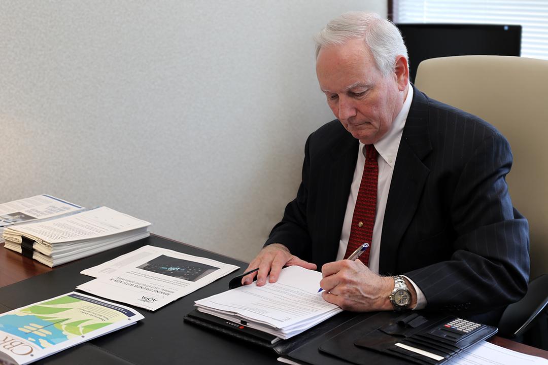A man in a suit and tie sits at a desk, reviewing papers with a focused expression.